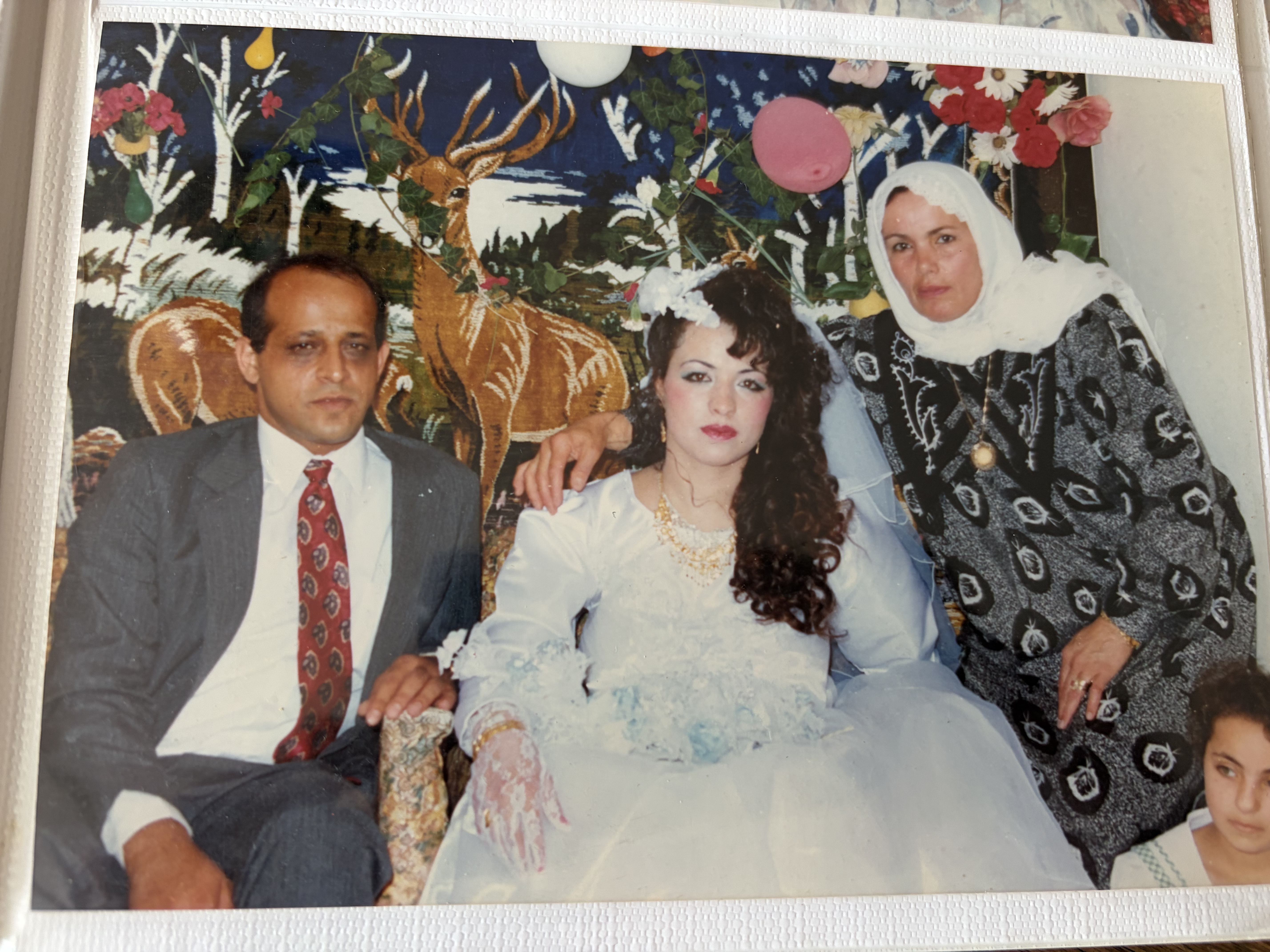 An old photograph showing a groom on the left and a bride on the right. an older woman with a head covering stands behind the bride